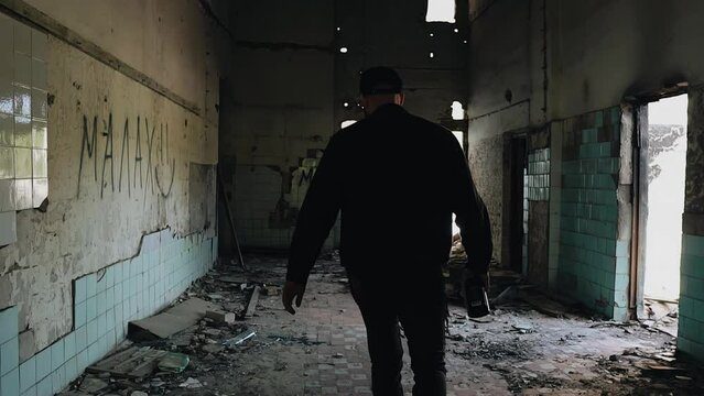 A drunk man with a bottle is walking along the corridor of an abandoned building. The camera shoots from behind