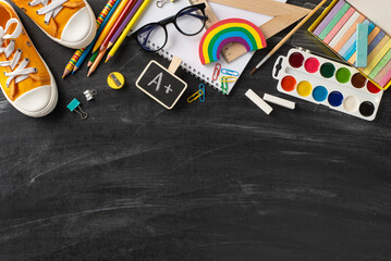 School supplies concept. Top view of colored pencils, art album, paint set, brush, A+ grade, ruler, clips, glasses, chalk, plasticine, sneakers and other on blackboard backdrop with empty area for ad