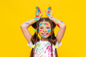 Portrait of a girl with a face painted with multicolored paints. Children's creativity. The child shows his ears above his head. Yellow isolated background.