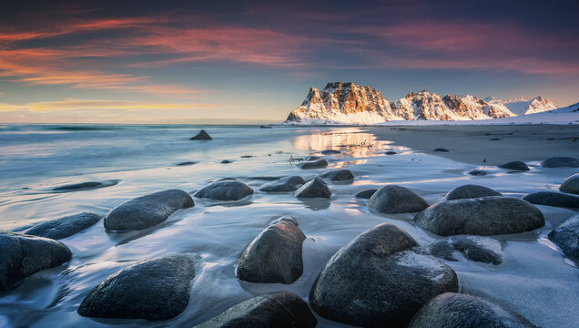 Rocky beach in winter during sunset. Magical north landscape. Sea coast with stones, blurred water, snowy rocky mountains, purple sky with clouds at dusk. Uttakleiv beach in Lofoten islands, Norway