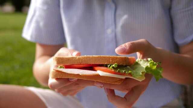 Close-up of a young girl sitting on the grass with a sandwich in her hands. A student eats a juicy sandwich in the campus park in sunny weather. 