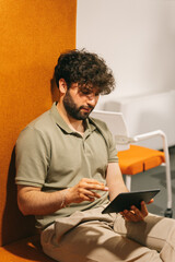 An attractive 30s male person working on digital tablet while enjoying lying on an orange sofa at the office