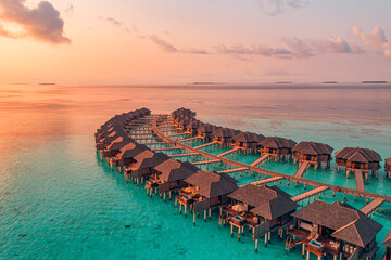 Sunset on Maldives island, luxury water villas resort and wooden pier. Beautiful aerial sky clouds and beach background. Summer coast vacation travel. Paradise sunrise landscape. Pristine sea bay - obrazy, fototapety, plakaty