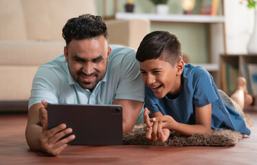 joyful indian father with son using digital tablet while lying on floor at home - concept of weekend holiday, Modern parenting and Digital entertainment