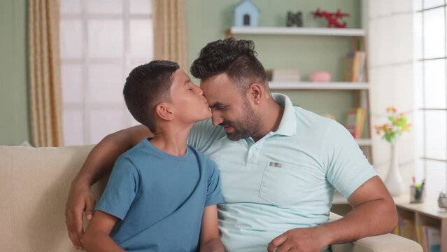 Happy indian son kissing his fathers forehead by looking at camera while sitting on sofa at home - concept of affection, Family love and tender moment.