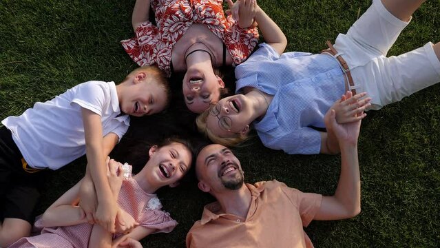 A big happy family lies in a circle on a green lawn in the park in summer and laughs. Brother tickles his sister. View from above.