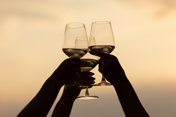 Silhouettes of hands of girls holding glasses of wine making a toast at the nature picnic on summer sunset.