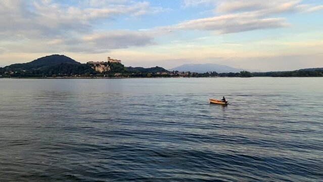 Fisherman rowing small wooden fishing boat on Maggiore lake in Italy with Angera fortress in background. Real-time