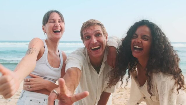 Young diverse friends laugh out loud and show thumbs up rejoicing at opportunity to have vacation together and visit tropical resort with warm sea stand on sandy beach on island. Multiethnic people