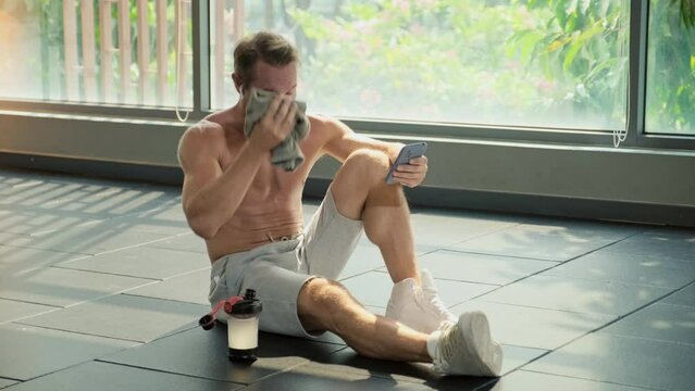 Long shot of sportsman sitting on floor, drinking water in bottle and serfing internet on smartphone after training sitting on floor in gym