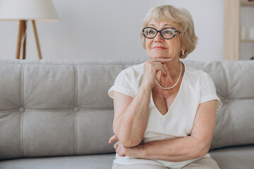 Smiling senior blonde haired woman looking at camera, happy old lady in glasses posing at home indoor, positive single senior retired female sitting on sofa in living room headshot portrait