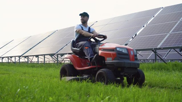 A man worker cutting grass with lawn mower on solar panels station