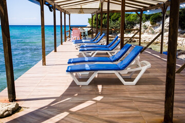 White sun loungers with blue cushions are waiting for their guests on the pier by the sea.