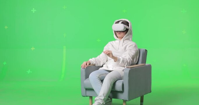 Horizontal shot on green screen background. VR or AR online at home. Cute girl in virtual reality headset in chair. Woman clicks invisible buttons and pulls virtual sliders. Beautiful woman have fun.