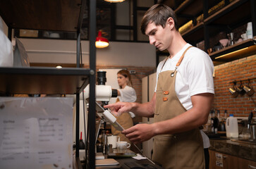 Portrait of a young male barista in apron using payment machine in cafe