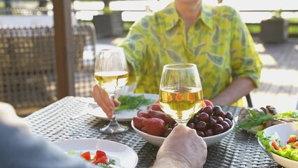 Close up of celebrating with glasses of wine or drink. Senior man and senior woman clinking glasses, having dinner outside. Eating, having fun together on weekend. Summertime on veranda. Slow motion