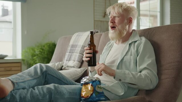 Leisure time of young bearded albino man, drinking beer, eating chips, sitting on sofa in living room in big modern house. Winner gesture. Football match. High quality 4k footage