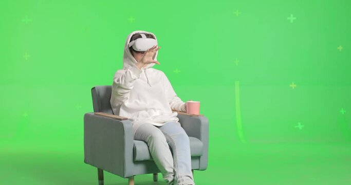 Shot on green screen background. VR or AR online at home. Cute girl in virtual reality googles in chair. Woman clicks invisible buttons pulls virtual sliders and drinking from a cup. Internet fun.