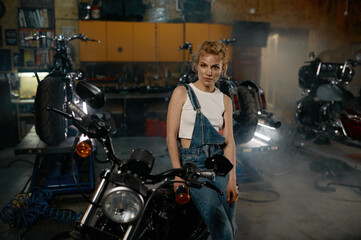 Fototapeta na wymiar Young woman in denim overalls looking at camera posing at motorcycle garage
