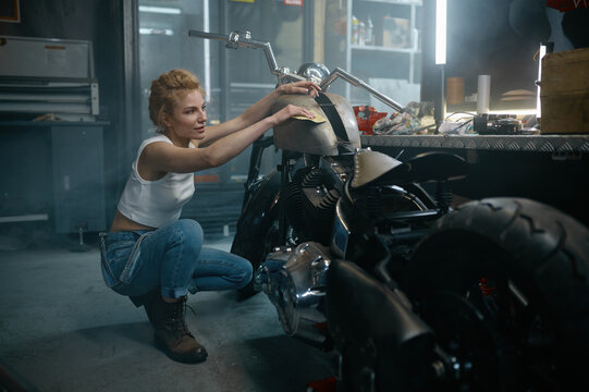 Young pretty woman mechanic polishing motorcycle after repair at workshop