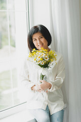 portrait of an asian woman with a bouquet of wild flowers at home near the window.