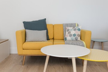 Bright and minimalist living room with comfortable cushions and gray blanket on a yellow sofa, round tables and a wooden floor
