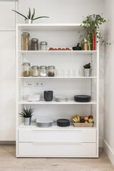Kitchenware, jars of food, books, fruit and plants on beautiful white shelving in a bright kitchen interior