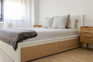 Interior of a bright minimalist bedroom with a wooden bed, plain sheets and a window with a white curtain