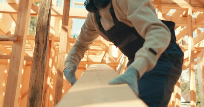 Builder man holding wooden plank, pulling it, strong man carrying board. Attractive guy in uniform builds house out of wood. Wooden house frame. Construction loader worker.