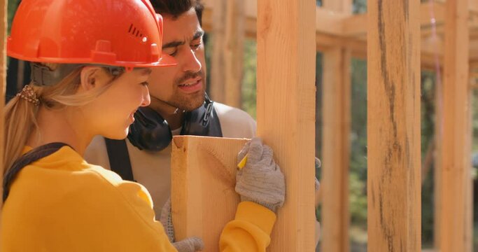 Man and woman workers in protective hardhat, helmet and headphones building wooden frame house on pile foundation. couple measuring wooden planks. Carpentry concept. Slow motion
