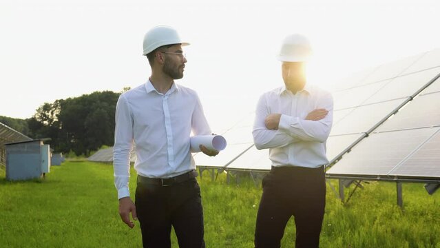 Two engineers walking at solar farm and discussing efficient plan of construction. Two men in protective helmet and uniform talking. Concept of alternative energy