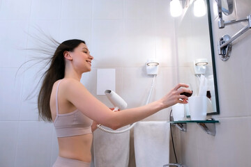 Beautiful brunette woman is looking at her reflection in the mirror and smiling while drying hair...