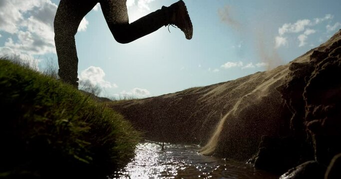 Young hikers actively run and jump through river water in wilderness. Travelers legs in trekking boots close up. Tourist lifestyle slow motion