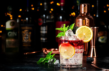 Bramble alcohol cocktail drink with dry gin, liqueur, syrup, lemon juice, blackberry and ice. Black bar counter background, steel bar tools and bottles - obrazy, fototapety, plakaty