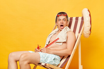 Shocked scared shirtless man sitting on deck chair isolated over yellow background looking nat camera with open mouth and big eyes.