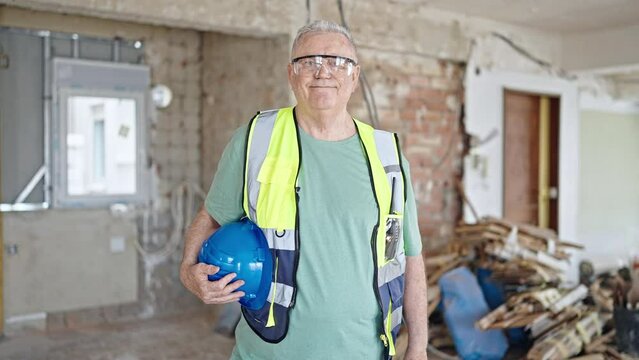 Middle age grey-haired man builder smiling confident holding hardhat at construction site
