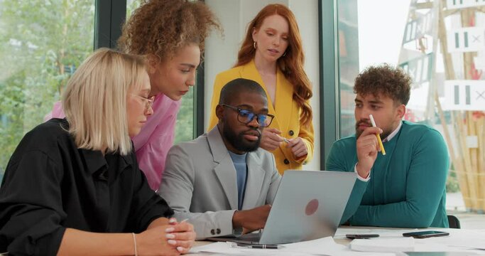bearded African American man explaining online job using laptop computer, talking, having teamwork discussion in office. Slow motion redhead woman holding sheet of papre asking advice