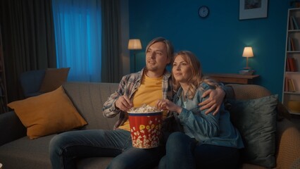 Obraz premium A young romantic couple is watching a movie and eating popcorn. Smiling man and woman hugging and relaxing on a comfortable sofa in the living room, enjoying a movie.