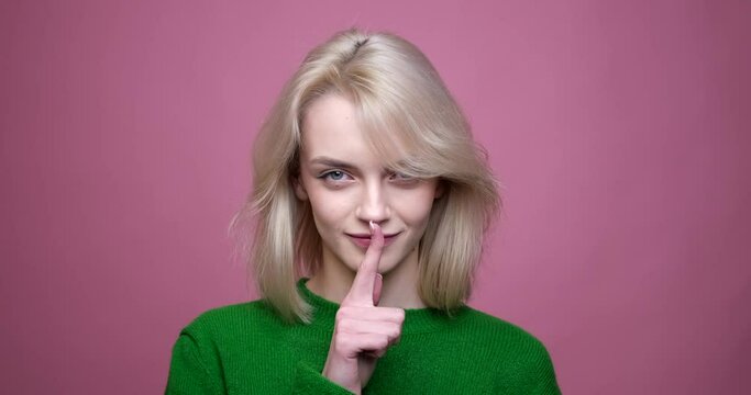 A Caucasian woman on a vibrant pink background. With a mischievous smile, she confidently raises her finger to her lips in a shushing gesture, accompanied by a playful wink.