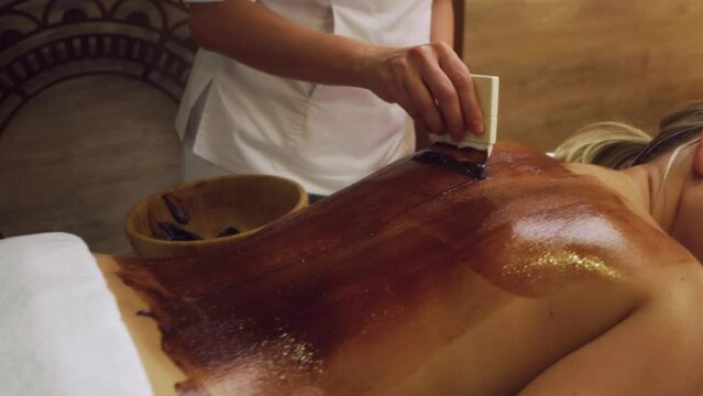 young slim fit woman having antioxidant back massage with chocolate body scrub in spa Slow motion, doctor covering patient's body with cosmetic chocolate.