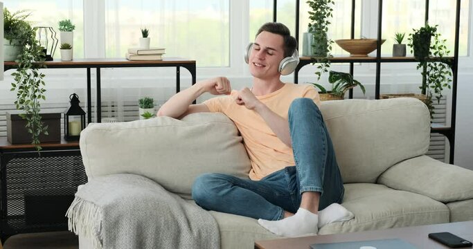 A young and cheerful Caucasian man is seated on a cozy couch in his home. He wears a pair of stylish headphones and is fully immersed in his music. A bright smile lights up his face.