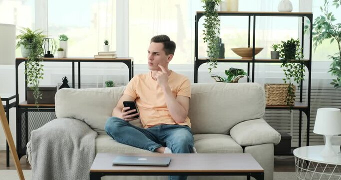 A thoughtful Caucasian man sits on a comfortable sofa, engrossed in his smartphone. With a pensive expression, he navigates through the digital realm, deep in contemplation.