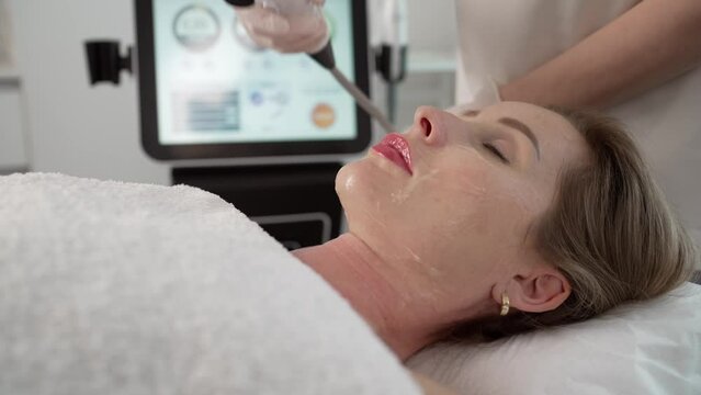 A dark-haired woman undergoes a skin rejuvenation procedure. A professional cosmetologist makes a procedure to a woman who lies on a couch. A middle-aged woman undergoes a skin rejuvenation procedure.