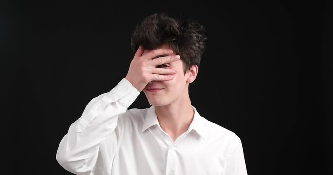 Charismatic Caucasian man engages in a mischievous act. With a sly smile on his face, he playfully covers his eyes with his hands, teasingly peering through his fingers.