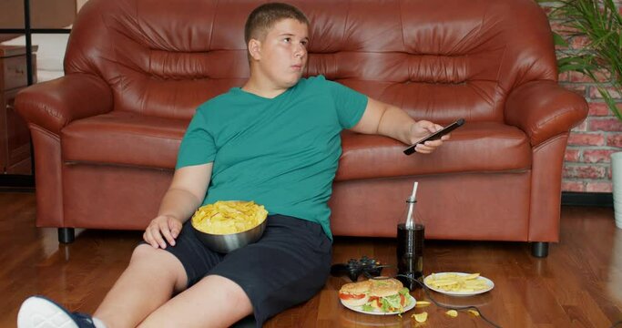 fat teenager boy is addicted to TV Slow motion overweight boy sit on sofa alone, has problems with nutrition, lead unhealthy lifestyle. laziness concept