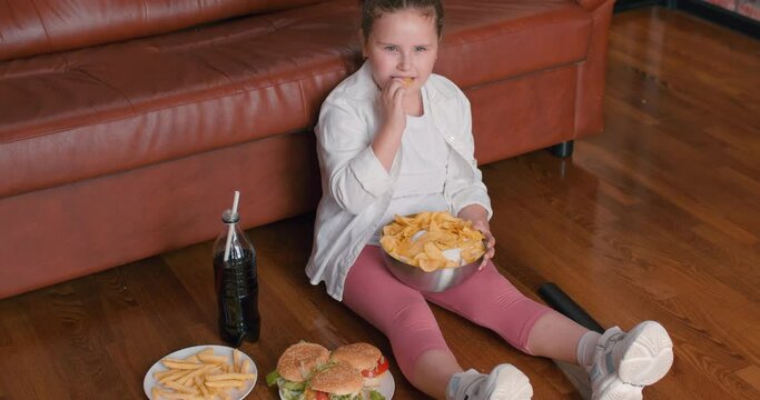 plump obese cute girl eating chips from bowl while sitting on floor in living room, Overweight caucasian child in casual clothes enjoy leading unhealthy lifestyle, eat junk food and watch tv.