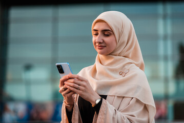 Portrait of a happy Muslim business woman in hijab using a phone near the office building.
