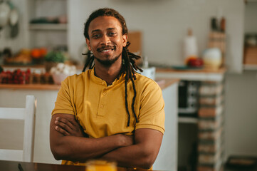 A cheerful interracial man is sitting at his cozy home with arms crossed and smiling at the camera.