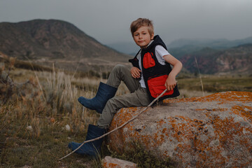 Boy hiker with stick in a vest and rubber boots sitting on the stone in the mountains