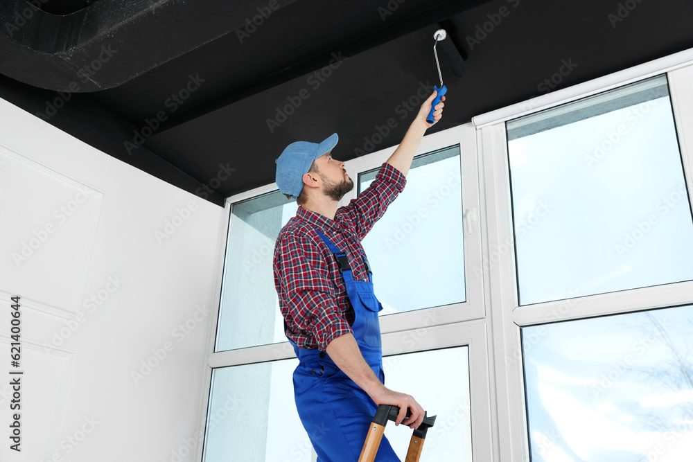 Wall mural worker in uniform painting ceiling with roller on stepladder indoors